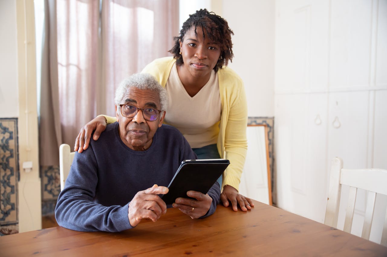 A senior man with eyeglasses and caregiver using a tablet at home, focusing on care and companionship.