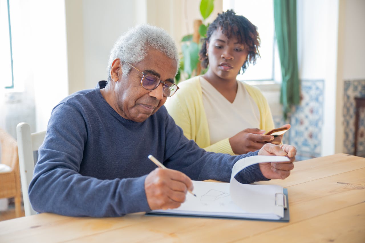 A senior man writing on papers with a caregiver nearby indoors in a supportive setting.